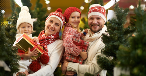 A cheerful family dressed in festive winter attire, holding gifts and standing among Christmas trees