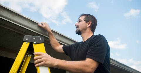 Home inspector on a ladder examining a roof and gutter system.