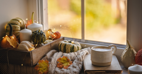 Cozy fall scene by a window featuring decorative pumpkins, candles, and a warm drink on a knitted blanket