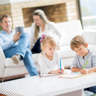 Two children doing homework at a table in a bright living room while parents relax in the background